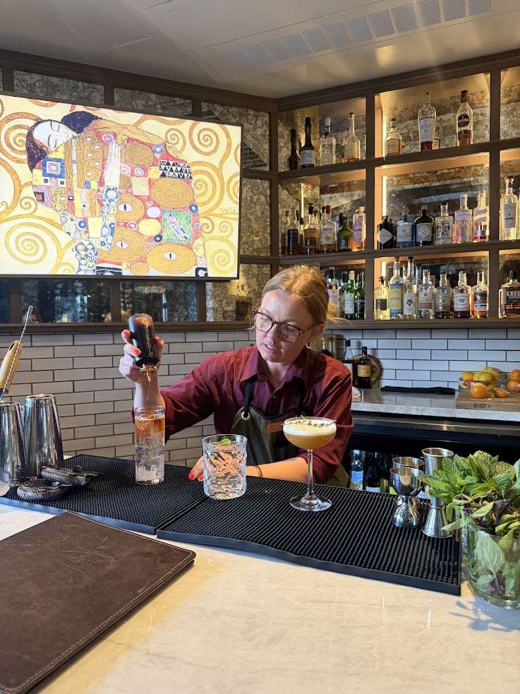 A bartender at the Rusty Parrot Lodge and Spa prepares a drink at the bar, smiling, with bottles and a cocktail on the counter.