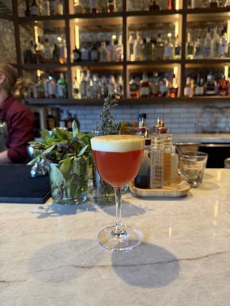 A cocktail with a frothy top sits on a marble bar at the Rusty Parrot Lodge and Spa, with bottles and herbs in the background.