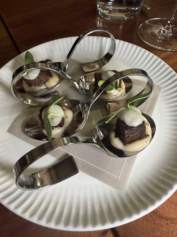 Four elegant metal spoons with gourmet bites, garnished with greens and arranged on a white plate—an exquisite presentation inspired by the refined culinary experience at Rusty Parrot Lodge and Spa.