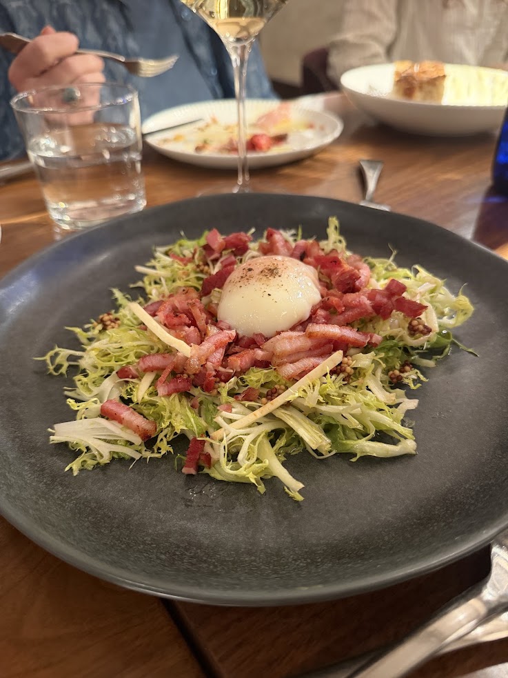 A salad with frisée, bacon, and a poached egg is served on a black plate at a restaurant table at the Rusty Parrot Lodge and Spa.