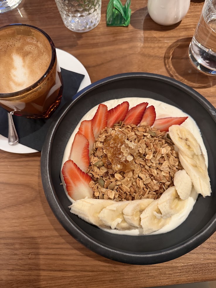 A bowl of yogurt with granola, strawberries, bananas, and honey sits beside a cup of coffee on a wooden table, evoking the cozy breakfast ambiance found at the Rusty Parrot Lodge and Spa.
