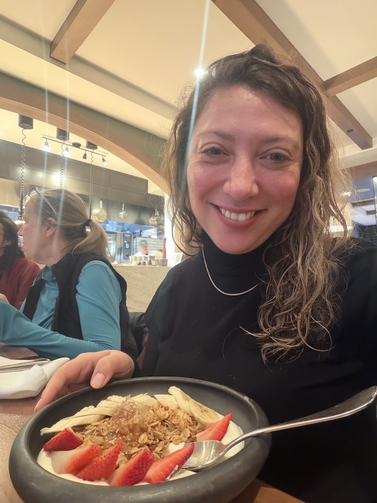 Smiling woman with curly hair holds a bowl of granola, bananas, and strawberries in a cozy restaurant at the Rusty Parrot Lodge and Spa.