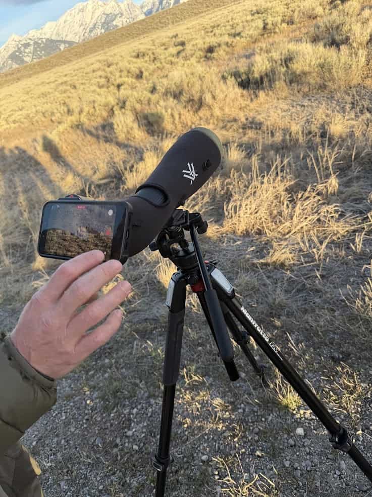 A hand holds a phone to a spotting scope on a tripod, aimed at a grassy hill with mountains in the background&mdash;a scene reminiscent of views found in any comprehensive Jackson Hole travel guide.