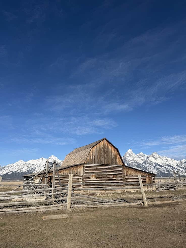 A wooden barn stands in front of snow-capped mountains under a clear blue sky, capturing the classic scenery featured in any Jackson Hole travel guide.