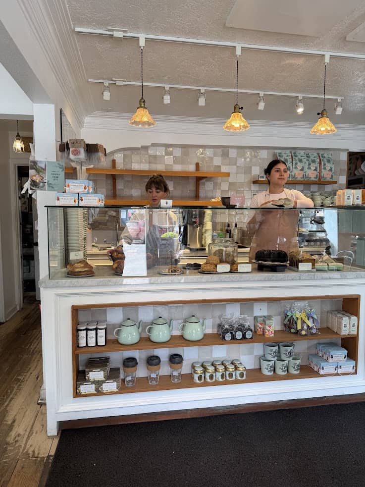 Two people stand behind a bakery counter with pastries, jars, and teapots on display in a bright shop&mdash;a charming scene you might find highlighted in any Jackson Hole travel guide.