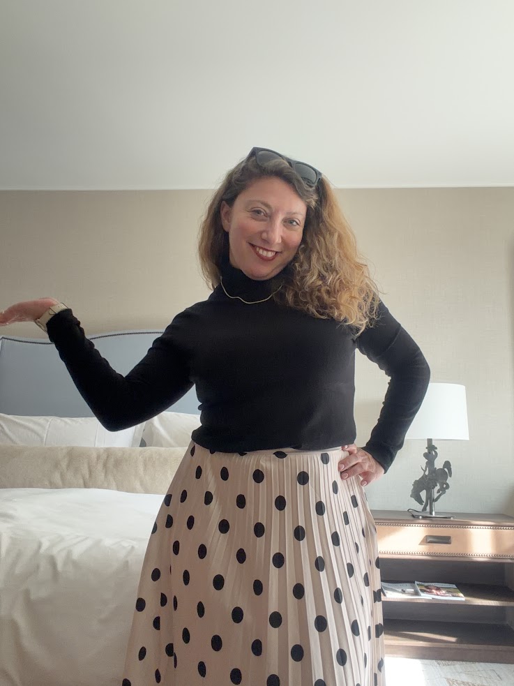 Woman in black top and polka dot skirt poses, smiling, in a well-lit bedroom at the Rusty Parrot Lodge and Spa, with a bed and lamp behind her.