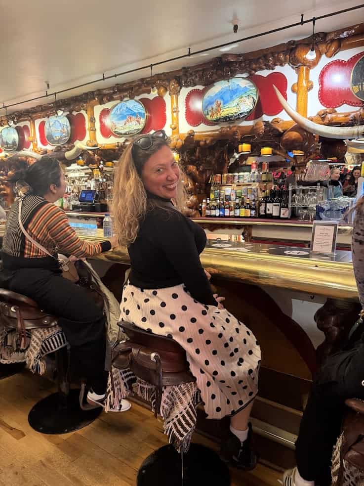 Woman in polka dot skirt smiles, sitting on a barstool at a colorful, western-themed bar&mdash;a moment right out of a Jackson Hole travel guide.
