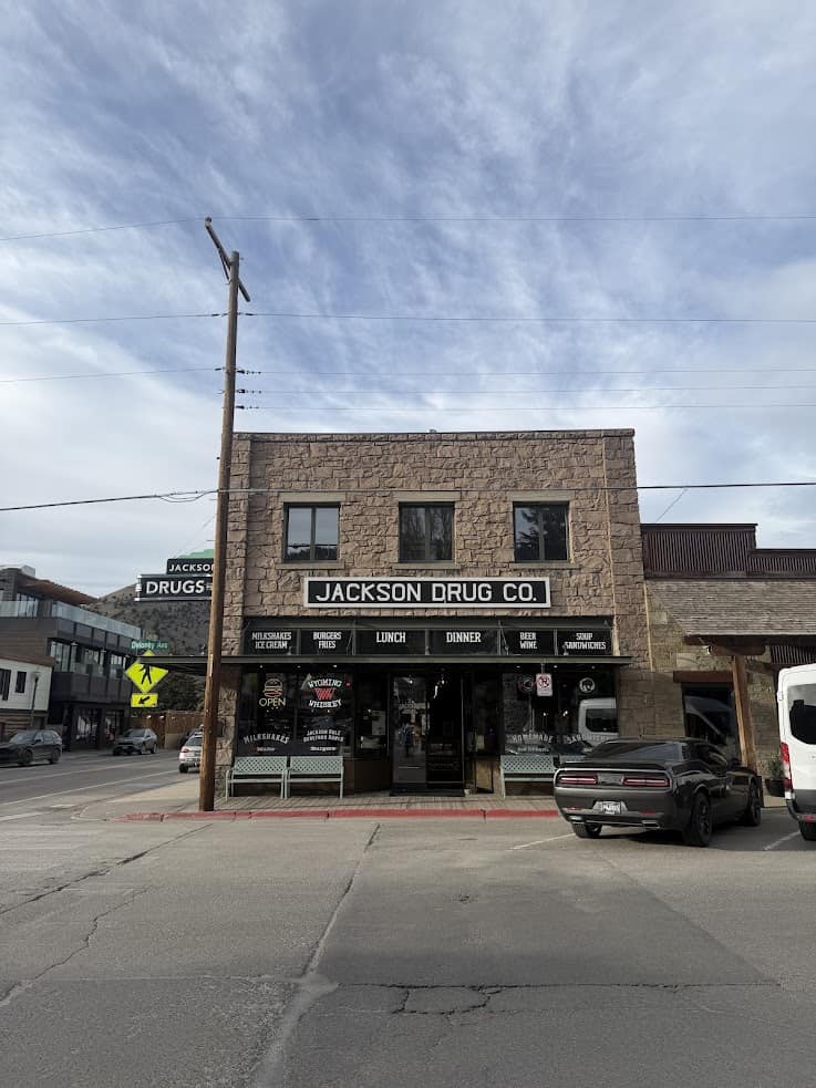 Two-story stone building with "Jackson Drug Co." signage, cars parked in front, and a clear sky above&mdash;an iconic stop often highlighted in any Jackson Hole travel guide.