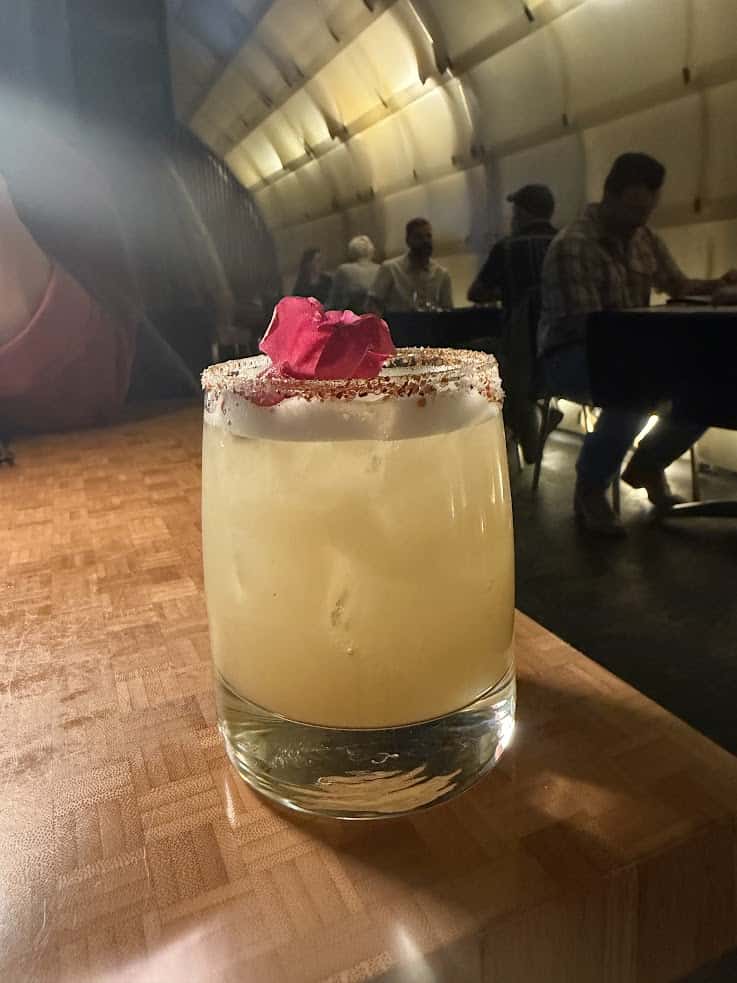 A yellow cocktail with ice, a chili-rimmed glass, and rose petals sits on a wooden table in a dim restaurant&mdash;an inviting scene you might find featured in an insider&rsquo;s Jackson Hole travel guide.
