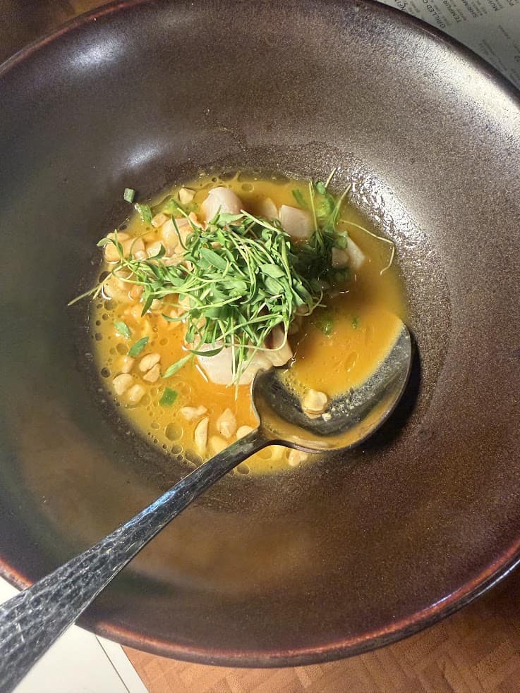 A bowl of soup with herbs, diced vegetables, peanuts, and a spoon resting on the side&mdash;a delicious comfort that&rsquo;s perfect after a day exploring with your Jackson Hole travel guide.