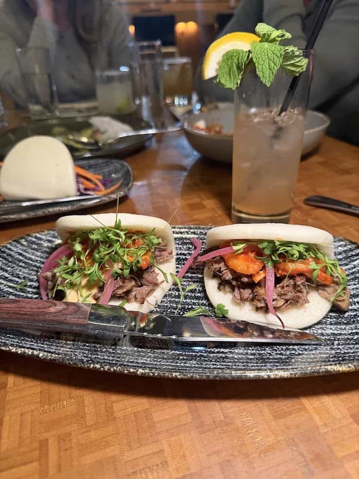 Two bao buns filled with meat, greens, and veggies on a plate with a knife&mdash;just the kind of delicious find you might discover in a jackson hole travel guide. A refreshing drink with lemon and mint sits behind, completing the scene.