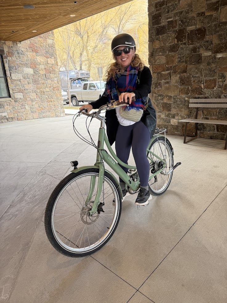 Smiling person wearing a helmet and scarf rides a green bicycle near a stone building, perhaps exploring like they&rsquo;ve found their own jackson hole travel guide to hidden gems.