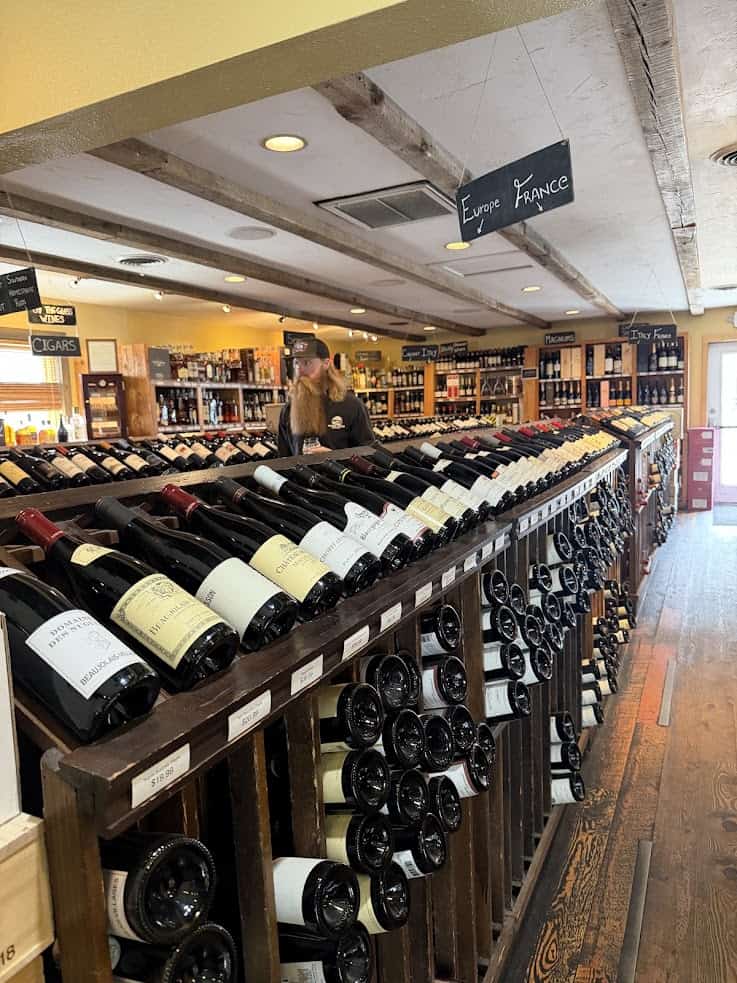 A man stands behind rows of wine bottles in a wine shop labeled &ldquo;Europe France,&rdquo; reminiscent of the charming discoveries one might find while following a detailed Jackson Hole travel guide, with wooden beams above adding to the rustic ambiance.