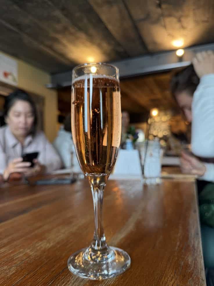 A glass of sparkling wine sits on a wooden table in a busy restaurant, with people blurred in the background&mdash;a scene reminiscent of the lively dining experiences highlighted in any great Jackson Hole travel guide.