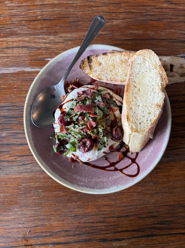 A plate with creamy dip topped with herbs and olives, served with two toasted bread slices and a spoon&mdash;just the kind of flavorful appetizer you might discover in a local eatery highlighted in a Jackson Hole travel guide.