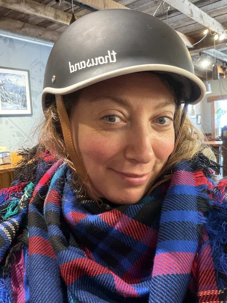 Woman wearing a black helmet and plaid scarf, smiling indoors with a rustic wooden ceiling overhead&mdash;capturing the cozy, adventurous spirit featured in any great Jackson Hole travel guide.