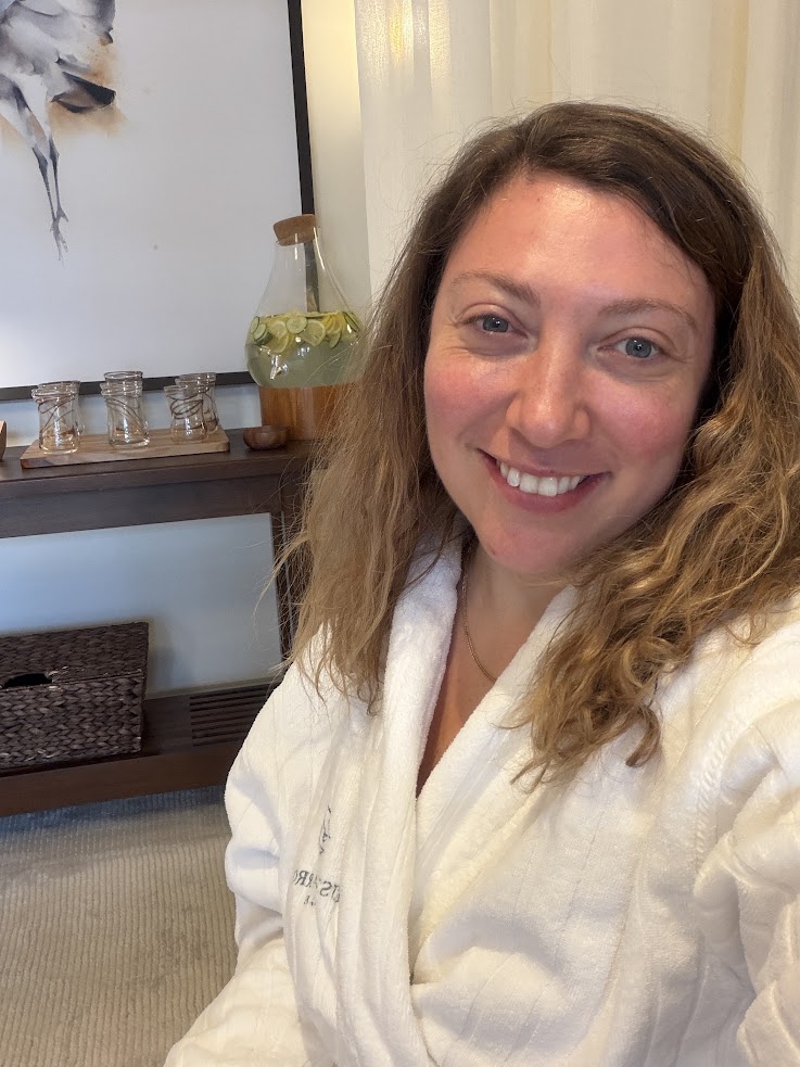 Woman in a white robe smiling indoors at the Rusty Parrot Lodge and Spa, with a drink dispenser and jars on a wooden shelf behind her.