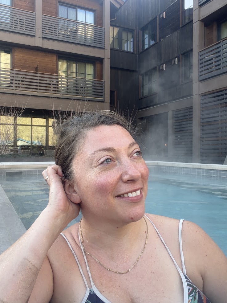 Woman smiling in an outdoor hot tub at the Rusty Parrot Lodge and Spa, with steam rising and a modern building in the background.