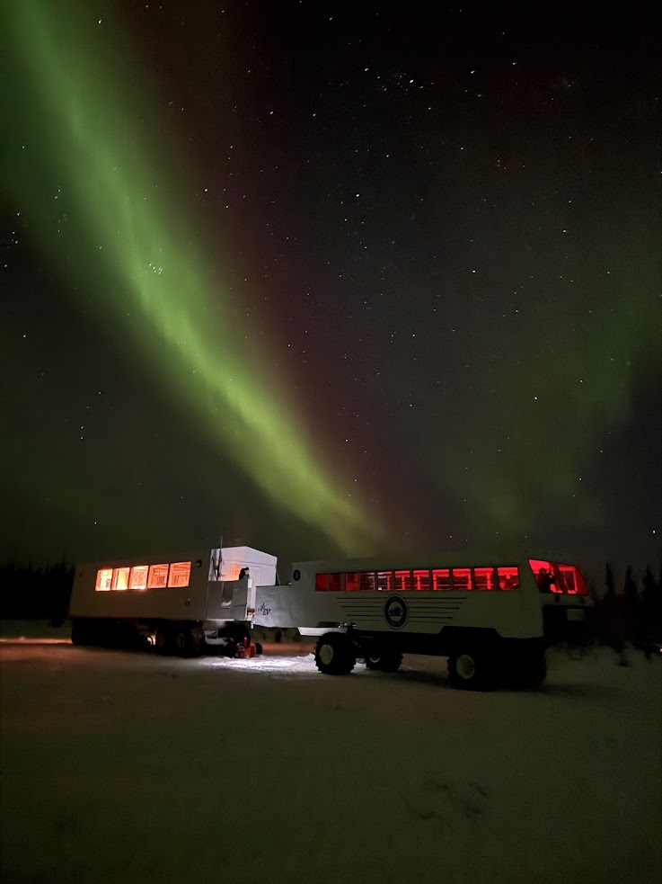 A vehicle with glowing windows under green northern lights in a snowy landscape at night, capturing the magical experience of a Churchill Northern Lights tour.