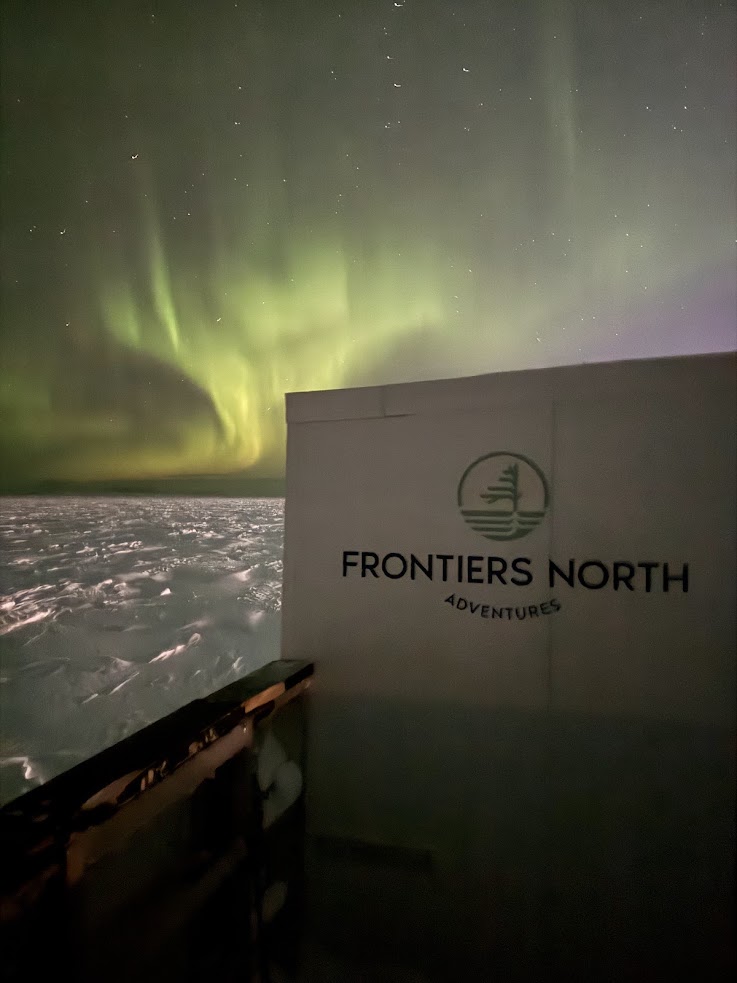 Green northern lights shimmer above snowy ground with a "Frontiers North Adventures" sign in the foreground at night, capturing the magic of a Churchill northern lights tour.
