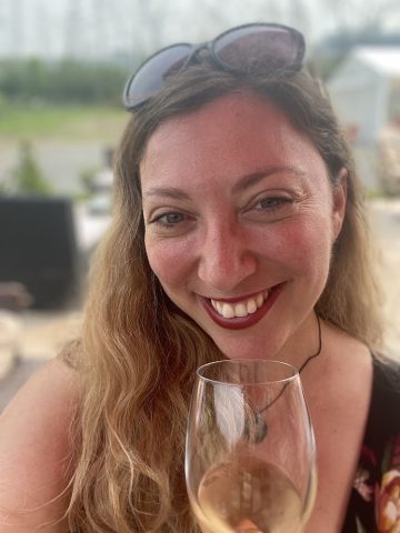 Smiling woman with long hair holding a glass of wine outdoors, enjoying wine tourism, sunglasses perched on her head.