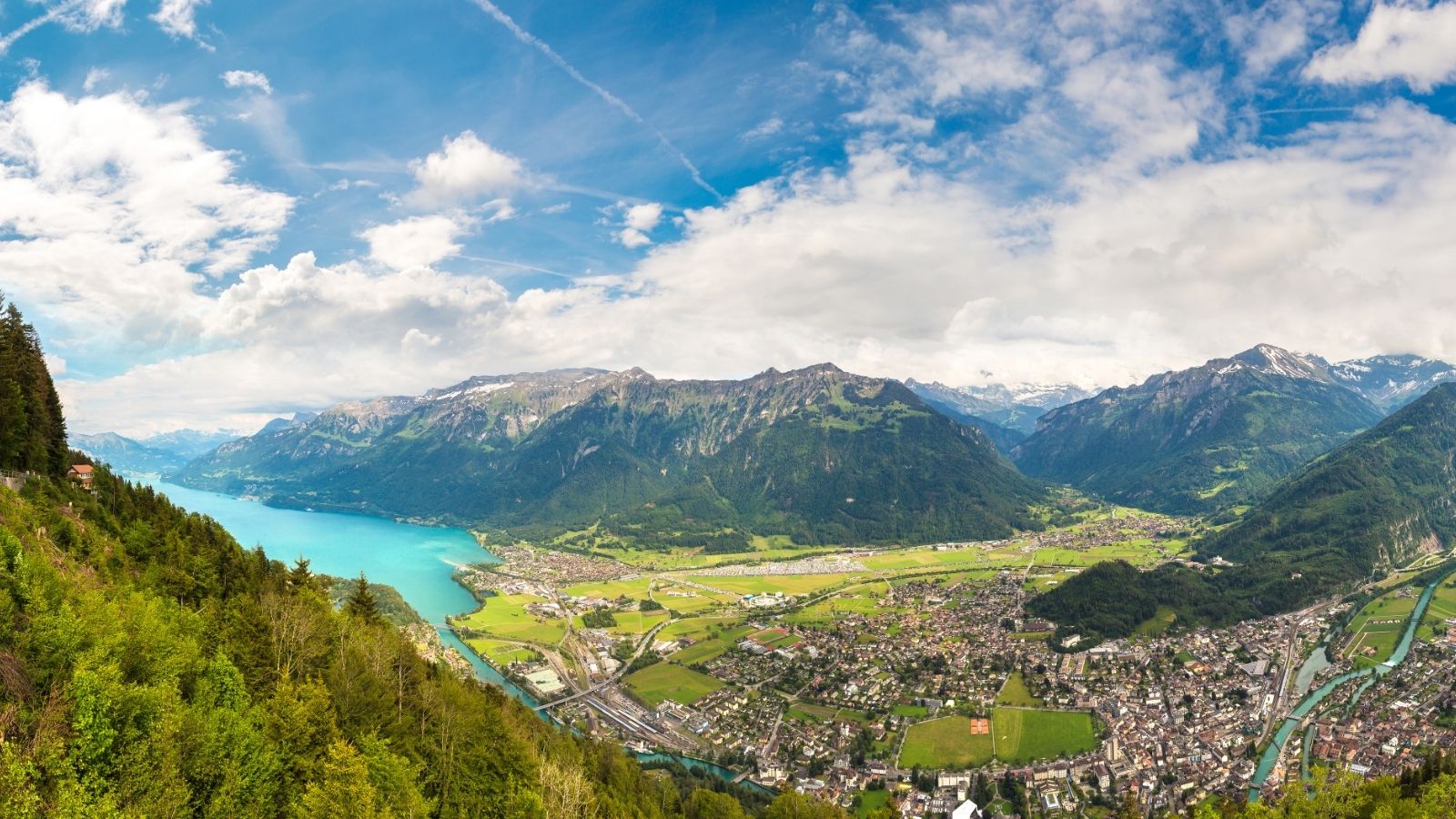 Aerial view of a town by a turquoise lake, surrounded by lush green mountains under a partly cloudy sky.