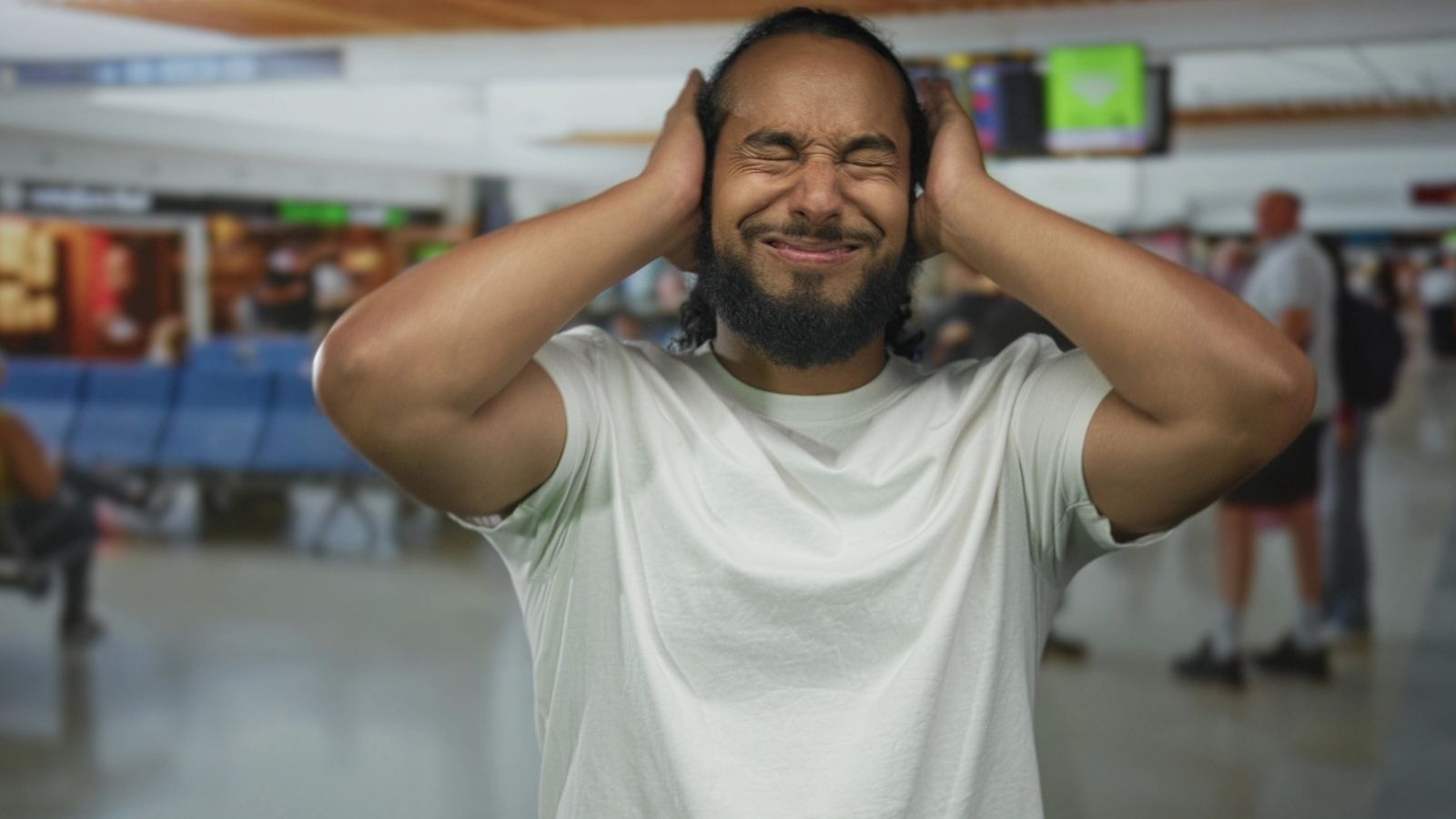 A man covers his ears and winces in discomfort in a busy airport terminal.