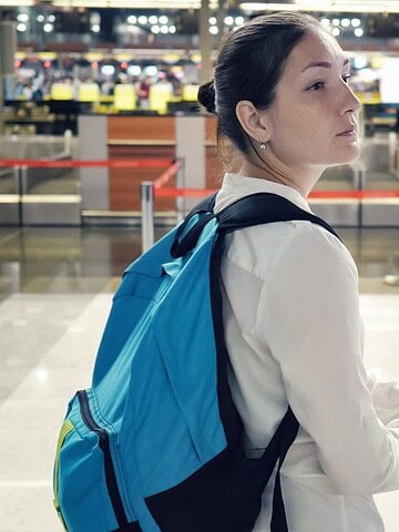 Woman with a blue backpack stands in an airport, holding luggage and looking to the side.