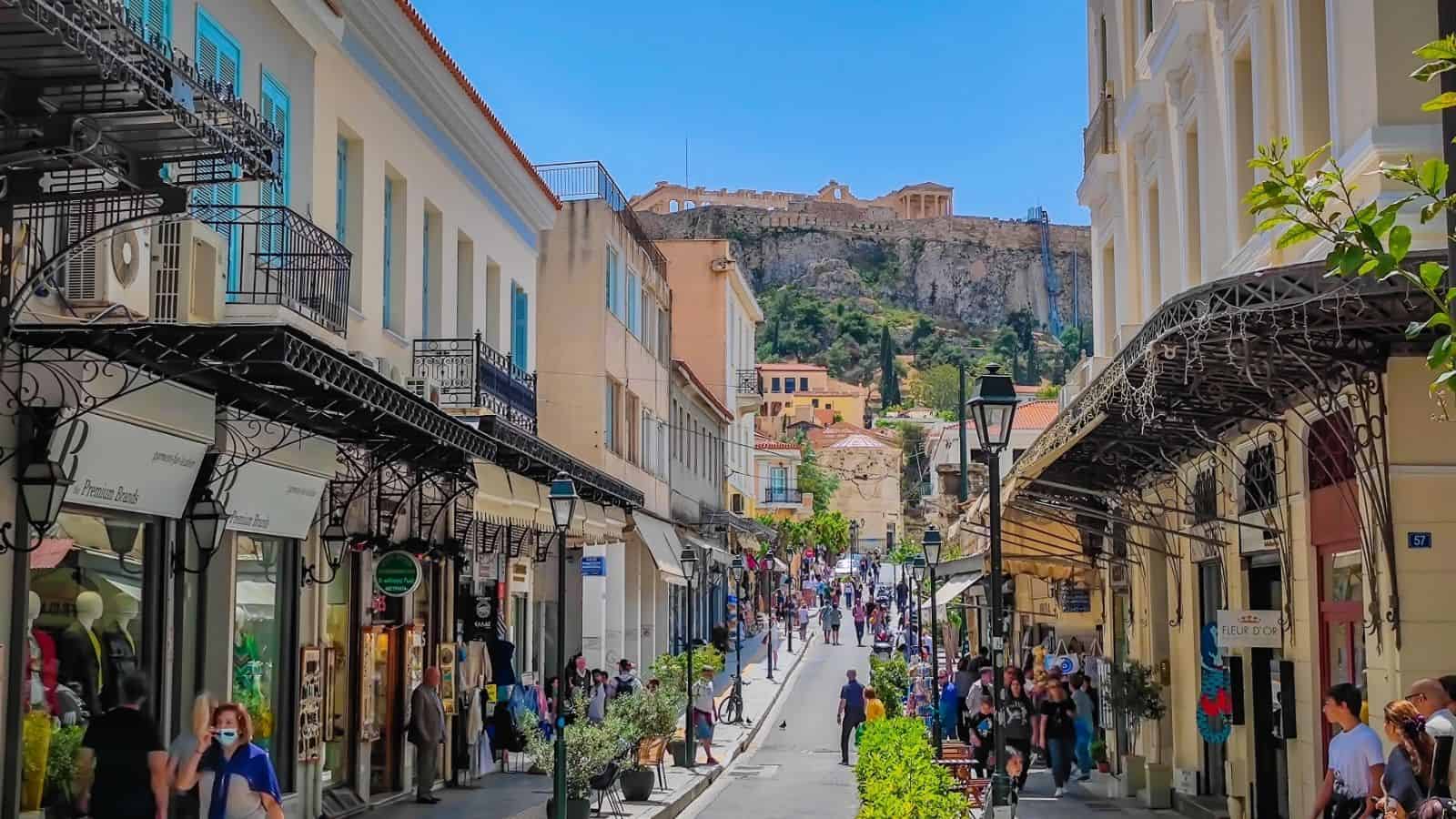 Busy street in Athens with shops, pedestrians, and the Acropolis visible in the background under a blue sky.