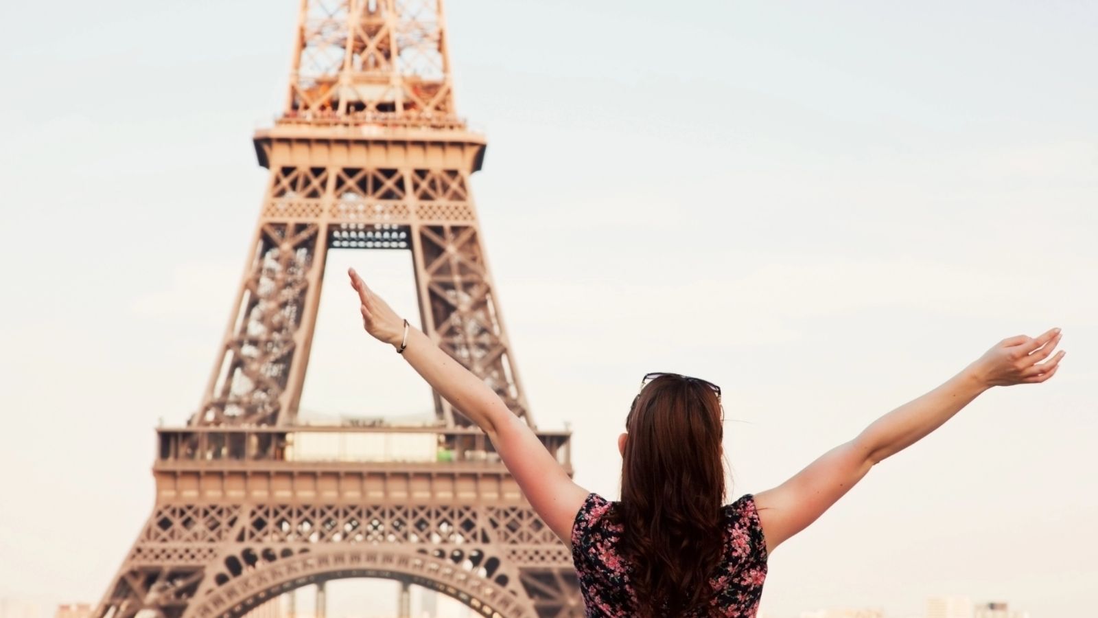 A woman with raised arms stands in front of the Eiffel Tower, facing away from the camera.