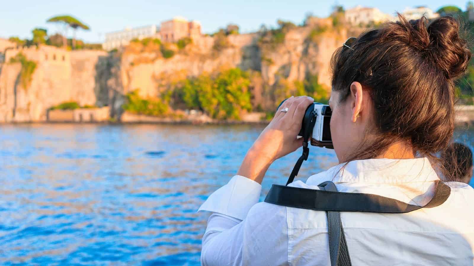 Woman taking a photo of cliffs and buildings across blue water, wearing a white shirt and camera strap. Perfect for those seeking scenic coastal destinations and easy to explore, car-free travel experiences.