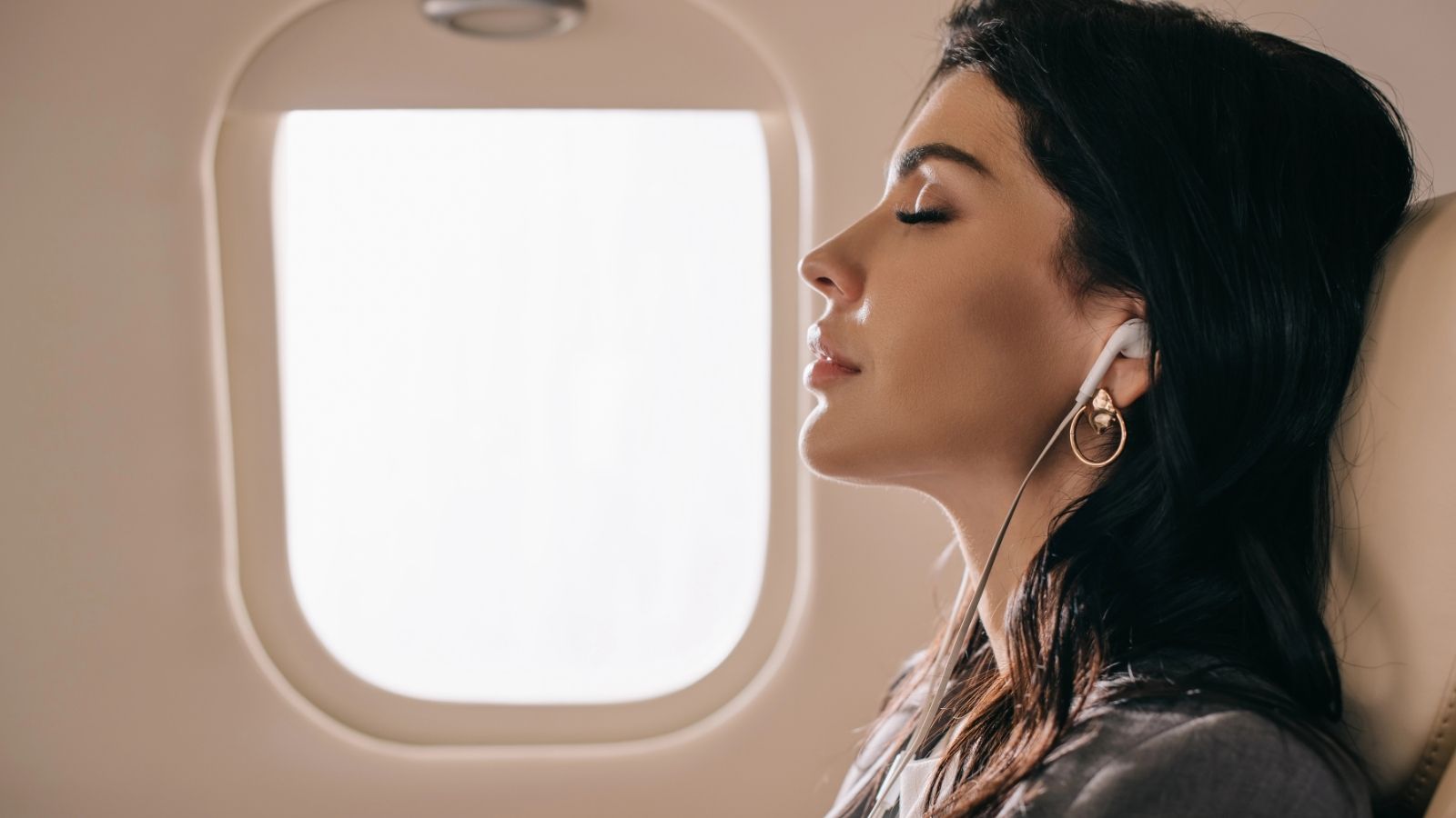 Woman with long dark hair, eyes closed, wearing earphones, relaxes in an airplane seat by the window.