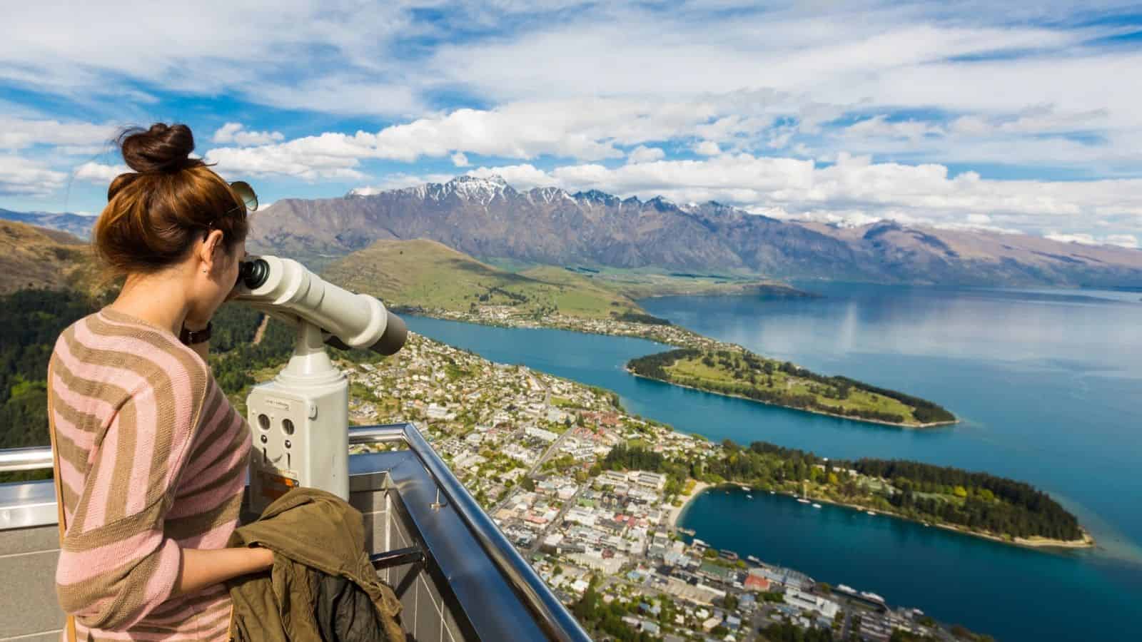Woman looks through a telescope at a scenic lake and mountain view from a high observation deck.