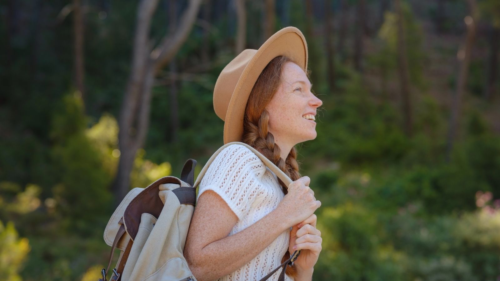 Smiling woman with a hat and backpack stands outdoors, surrounded by green trees and sunlight.