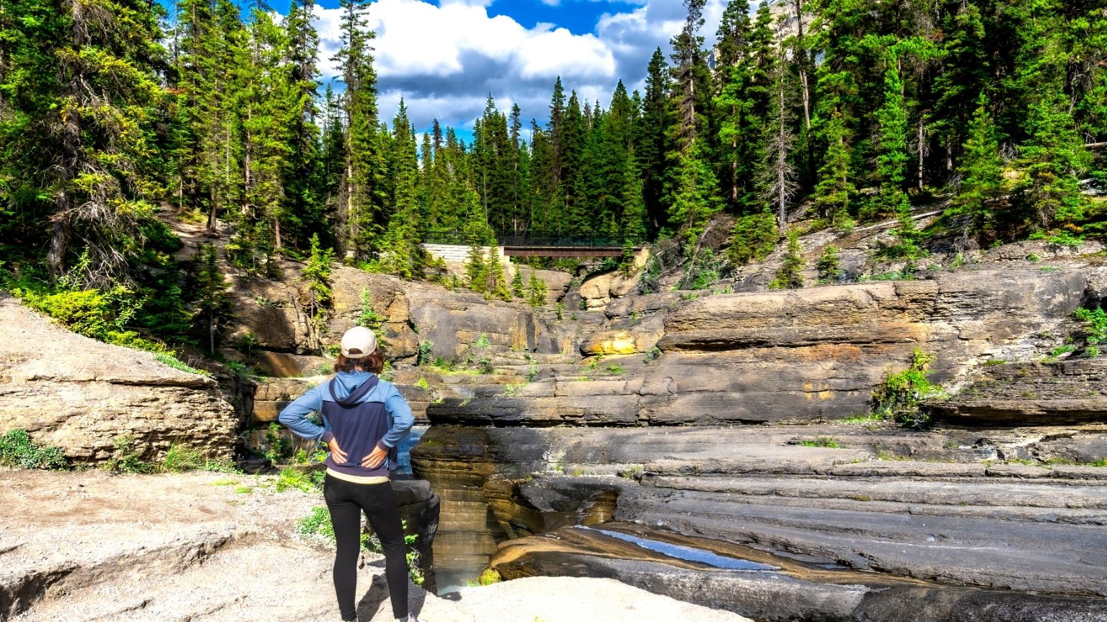 Person in a cap stands on rocky terrain, looking at forest and bridge under a blue sky with clouds.