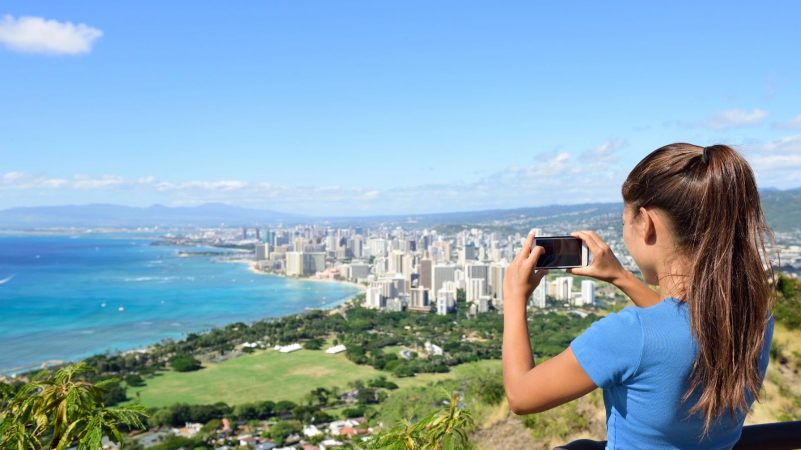 Girl taking a photo of a city skyline and ocean from a scenic viewpoint on a sunny day.