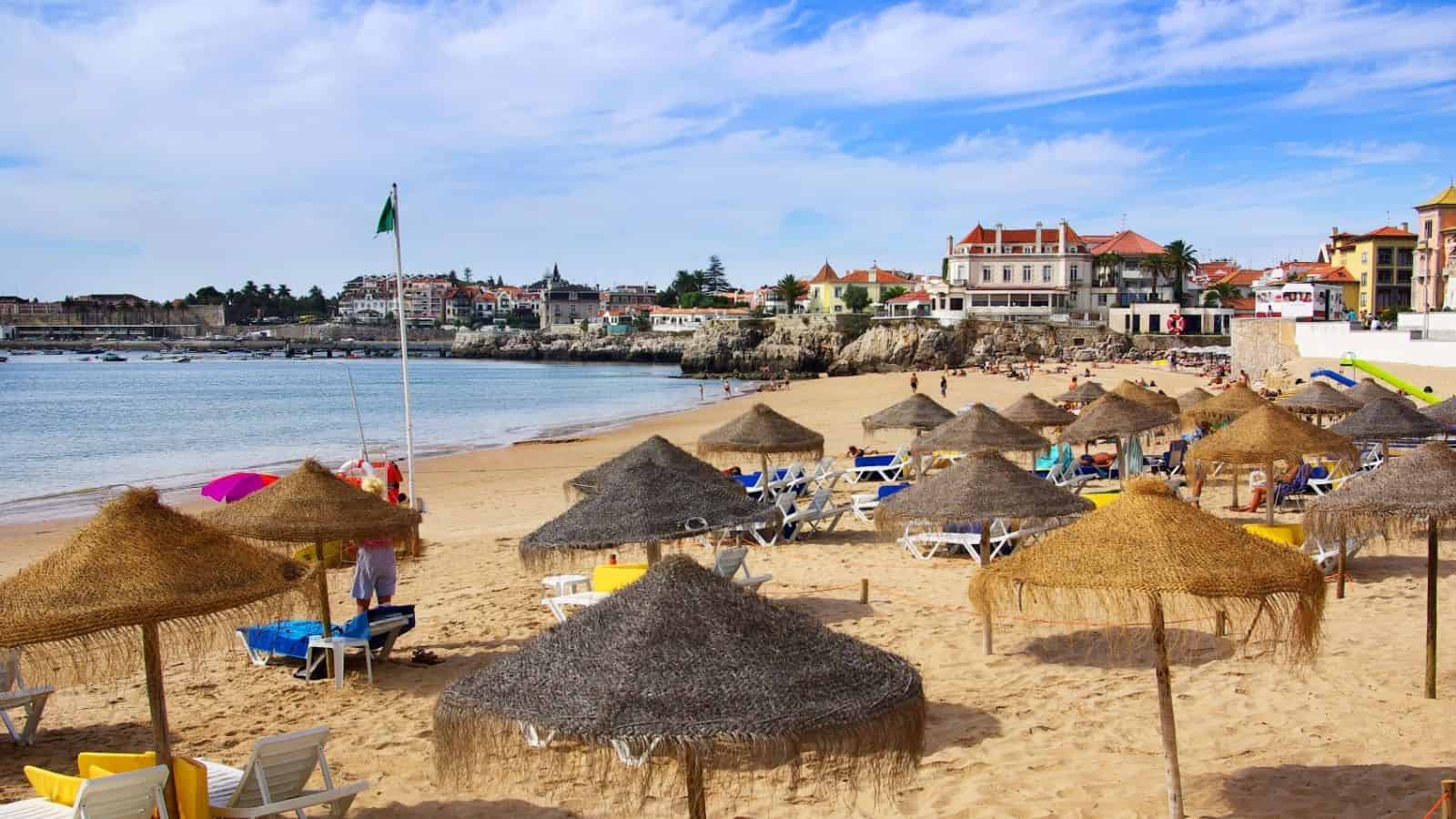 Straw umbrellas and lounge chairs on a sandy beach with buildings and blue sky in the background.