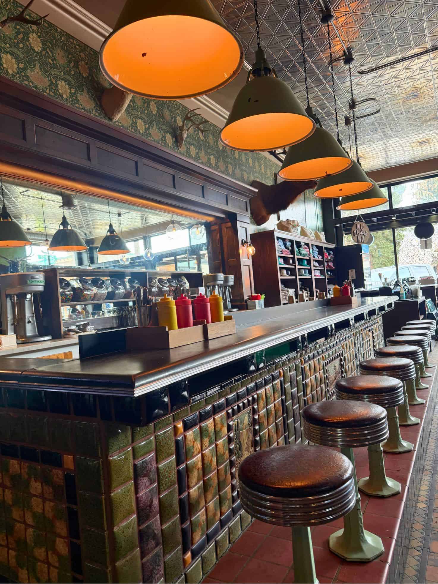 A retro diner counter with stools, condiment bottles, and hanging green lamps in a vintage setting&mdash;a scene reminiscent of classic Americana you might find highlighted in a Jackson Hole travel guide.