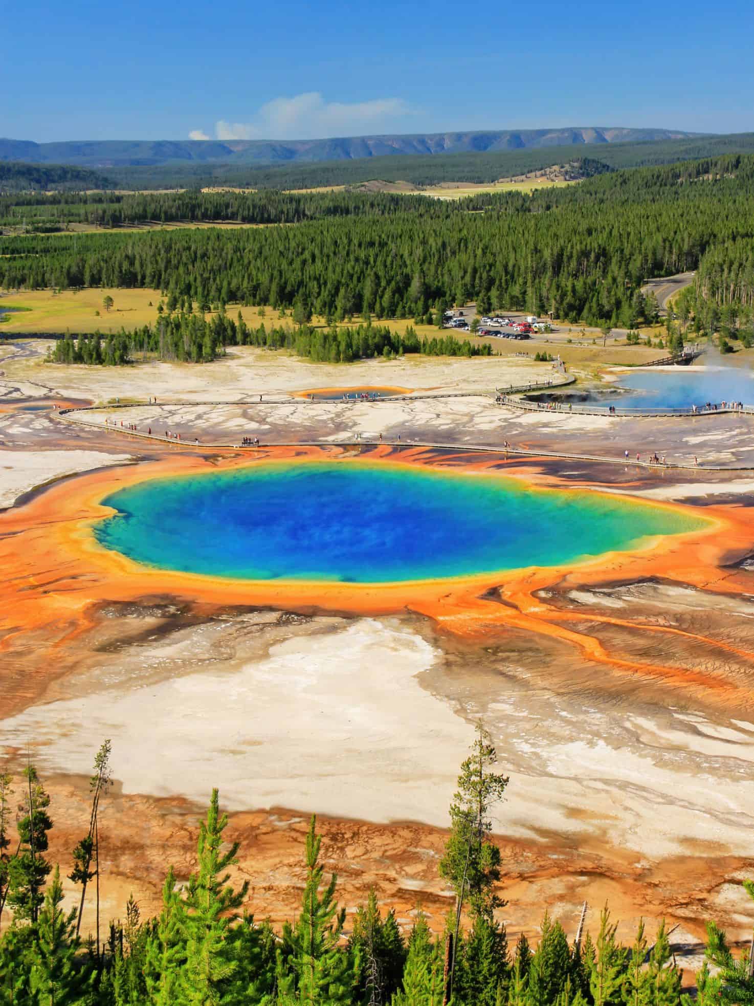 A vibrant hot spring with blue, green, yellow, and orange rings surrounded by trees and distant hills&mdash;a must-see highlighted in any Jackson Hole travel guide.
