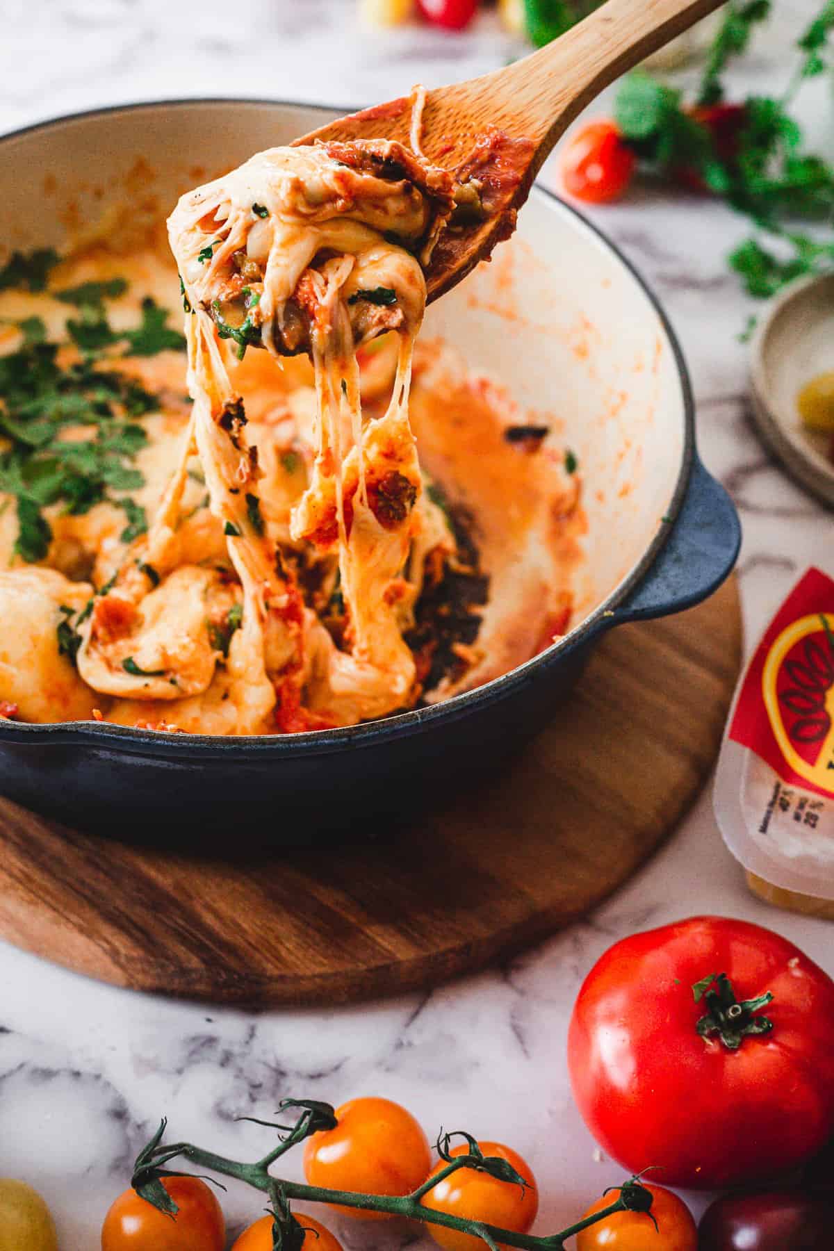 A wooden spoon lifts gooey, melted cheese from a tomato-based dish in a blue pot—an irresistible moment for those seeking foodie destinations, with fresh tomatoes completing the scene.