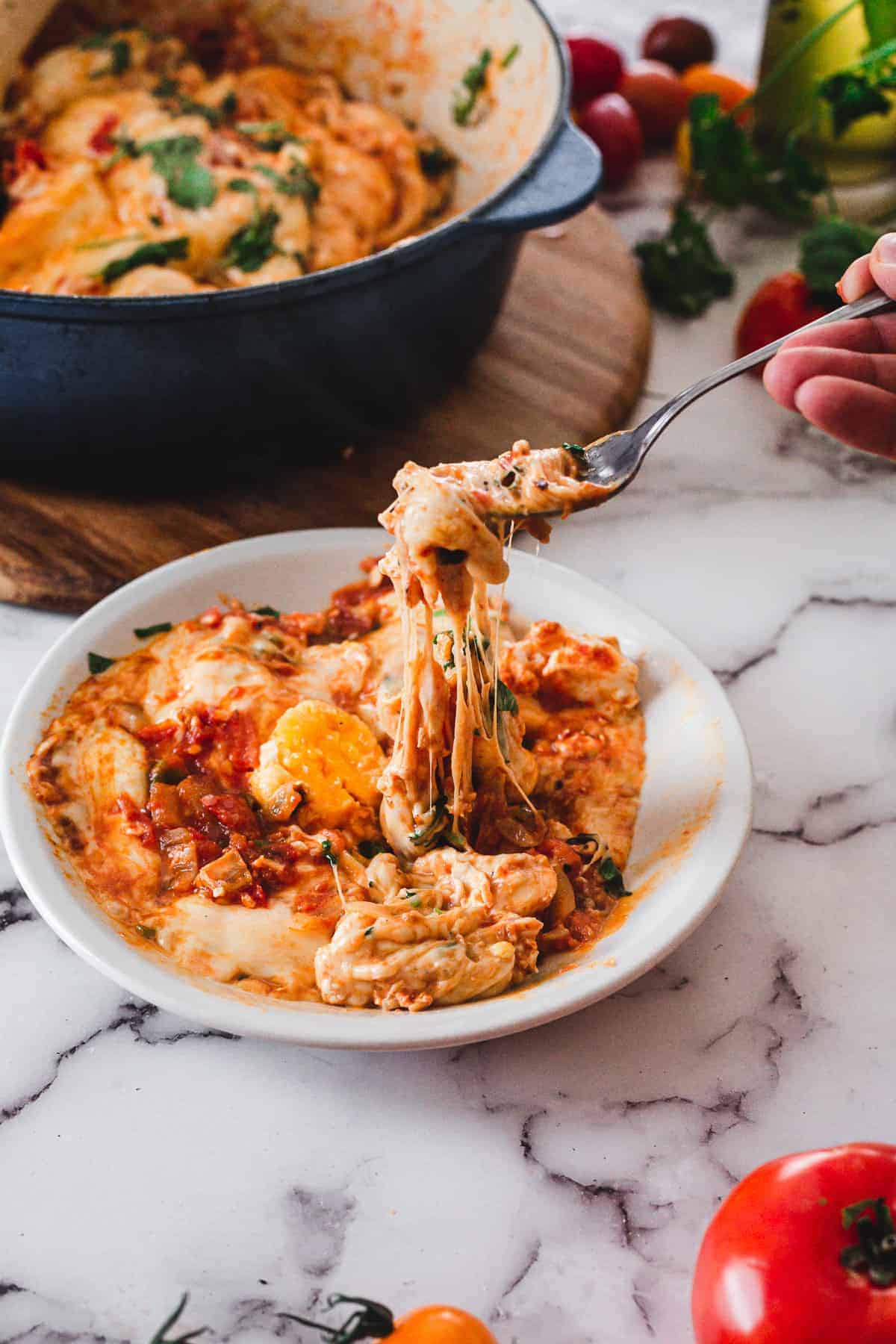 A fork lifts cheesy, saucy eggs from a plate, with a pot of cooked eggs and tomatoes in the background—a tempting scene straight from top foodie destinations.