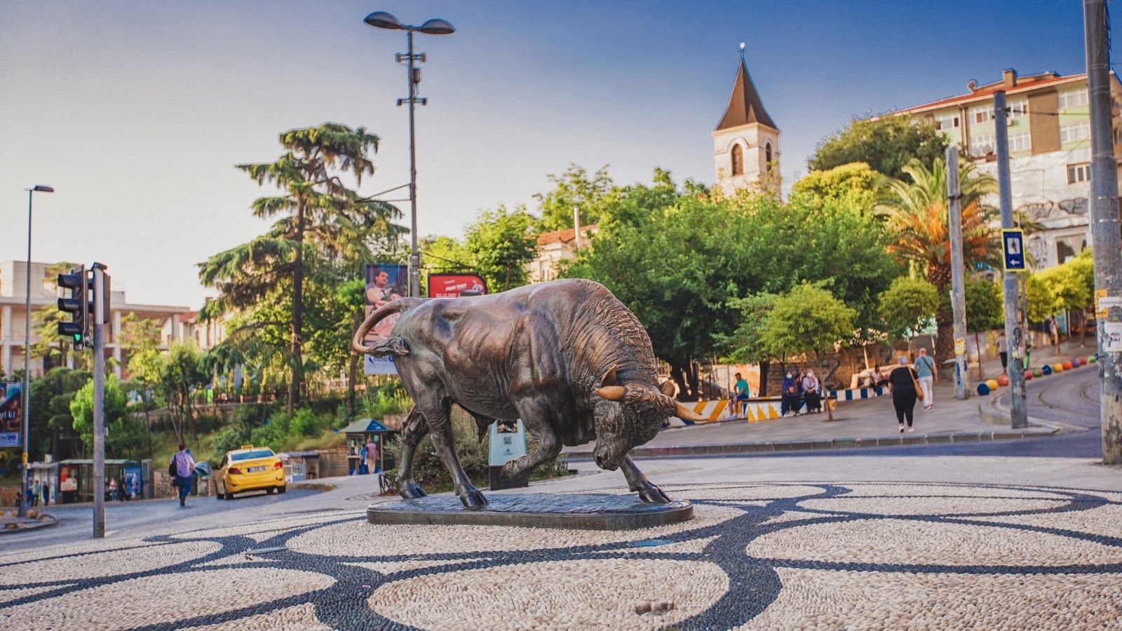 A bronze bull statue stands in a city plaza, surrounded by trees, buildings, and people walking.