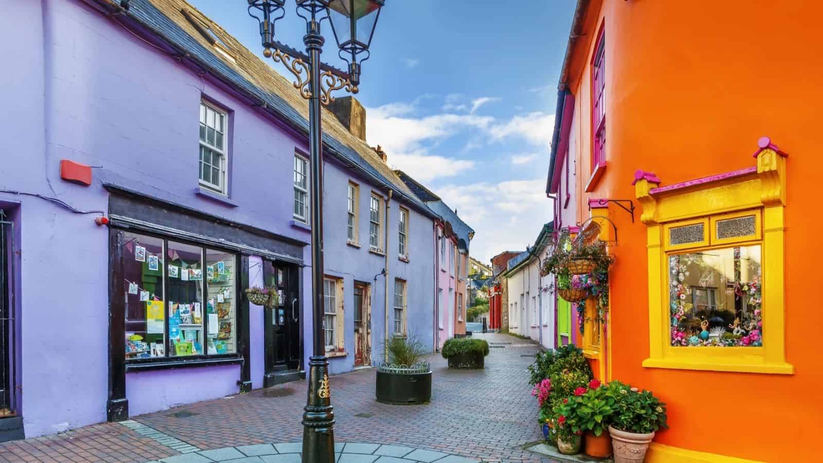 Colorful buildings line a narrow, cobbled street with flowers and a vintage lamp post under a blue sky.