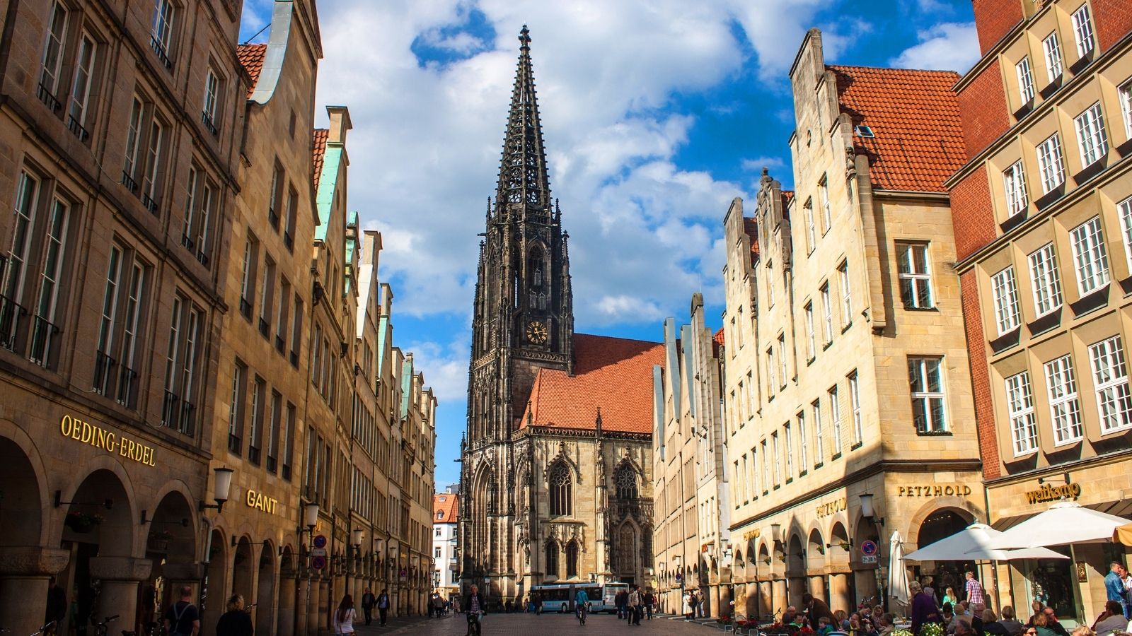 A tall Gothic church stands between historic buildings on a busy pedestrian street under a blue sky.
