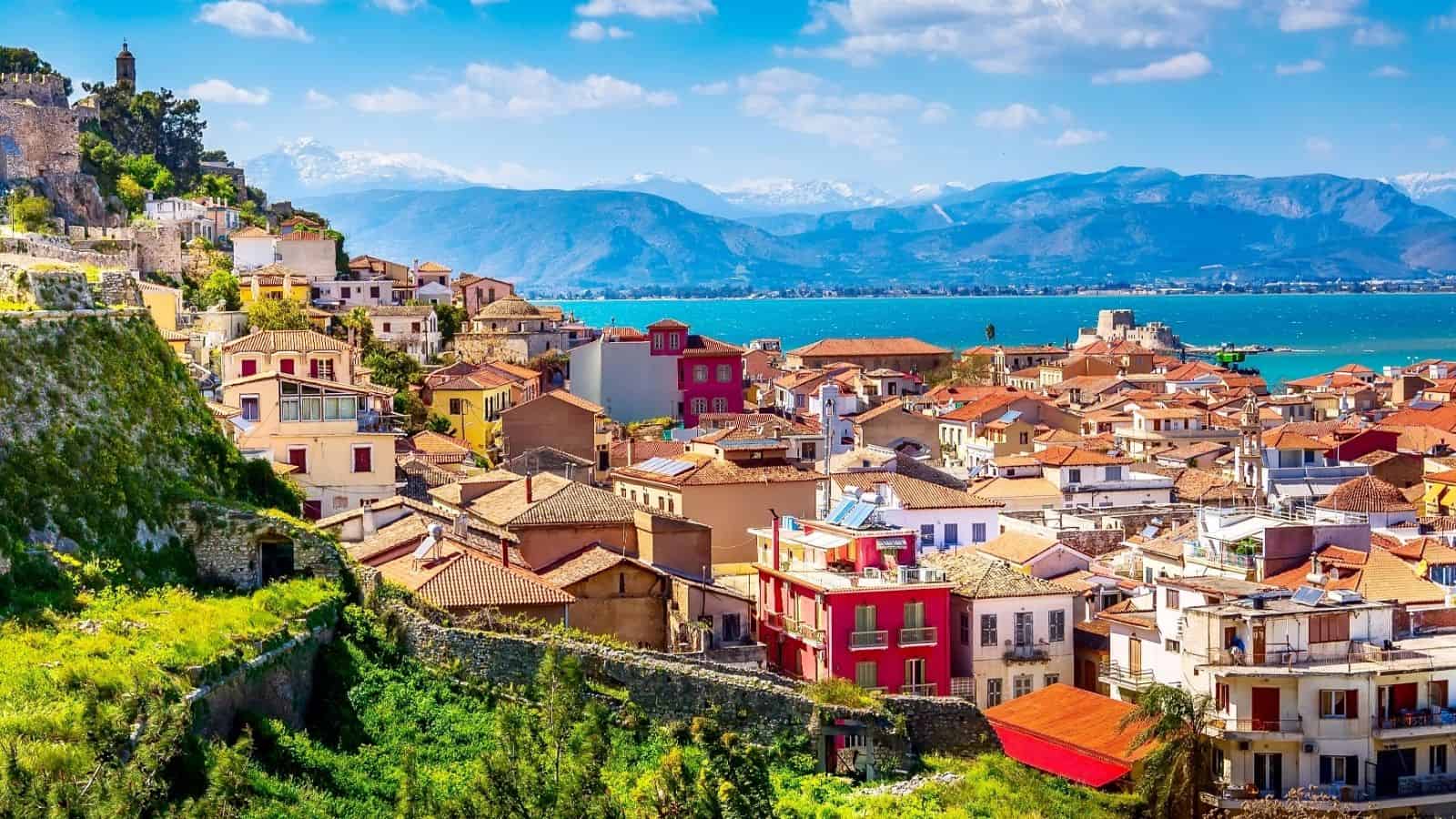 Colorful seaside town with red-roofed buildings, lush greenery, and mountains in the background.
