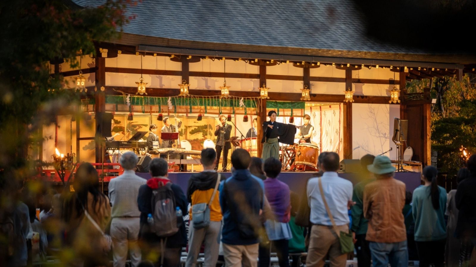 A crowd watches musicians perform on a stage at a traditional outdoor event in the evening.