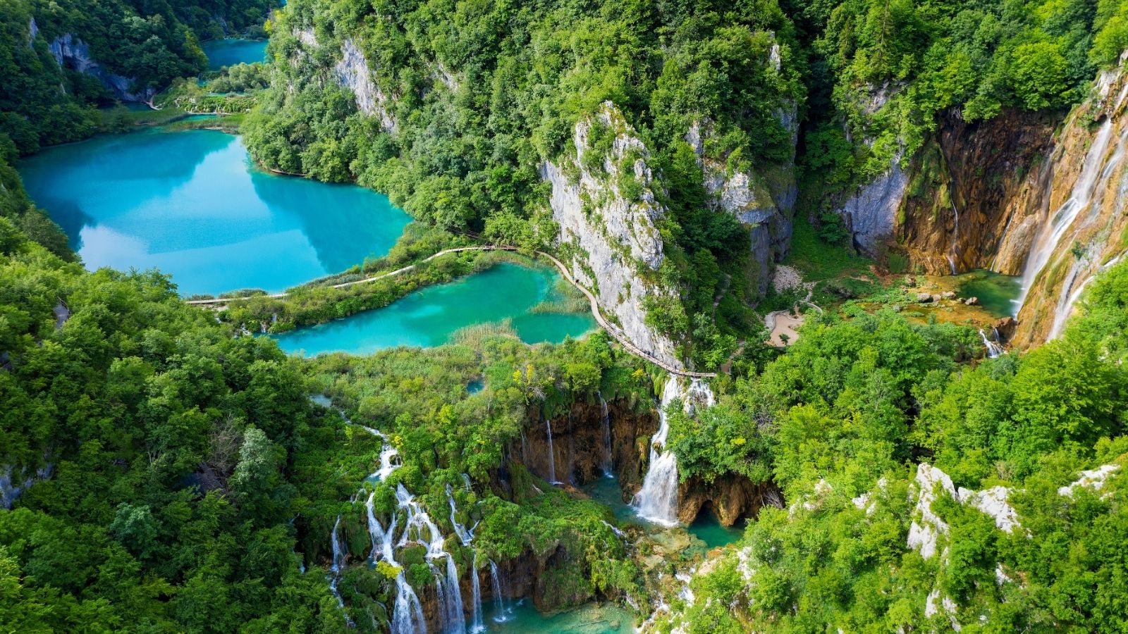 Aerial view of turquoise lakes, waterfalls, and lush green forest in a mountainous landscape.