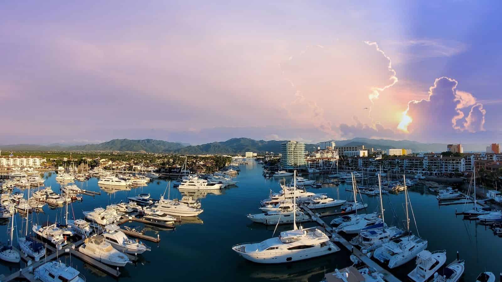 Boats and yachts docked at a marina with buildings, mountains, and a colorful sunset sky in the background, creating one of the most scenic coastal destinations that’s easy to explore on foot thanks to car-free travel along the waterfront.