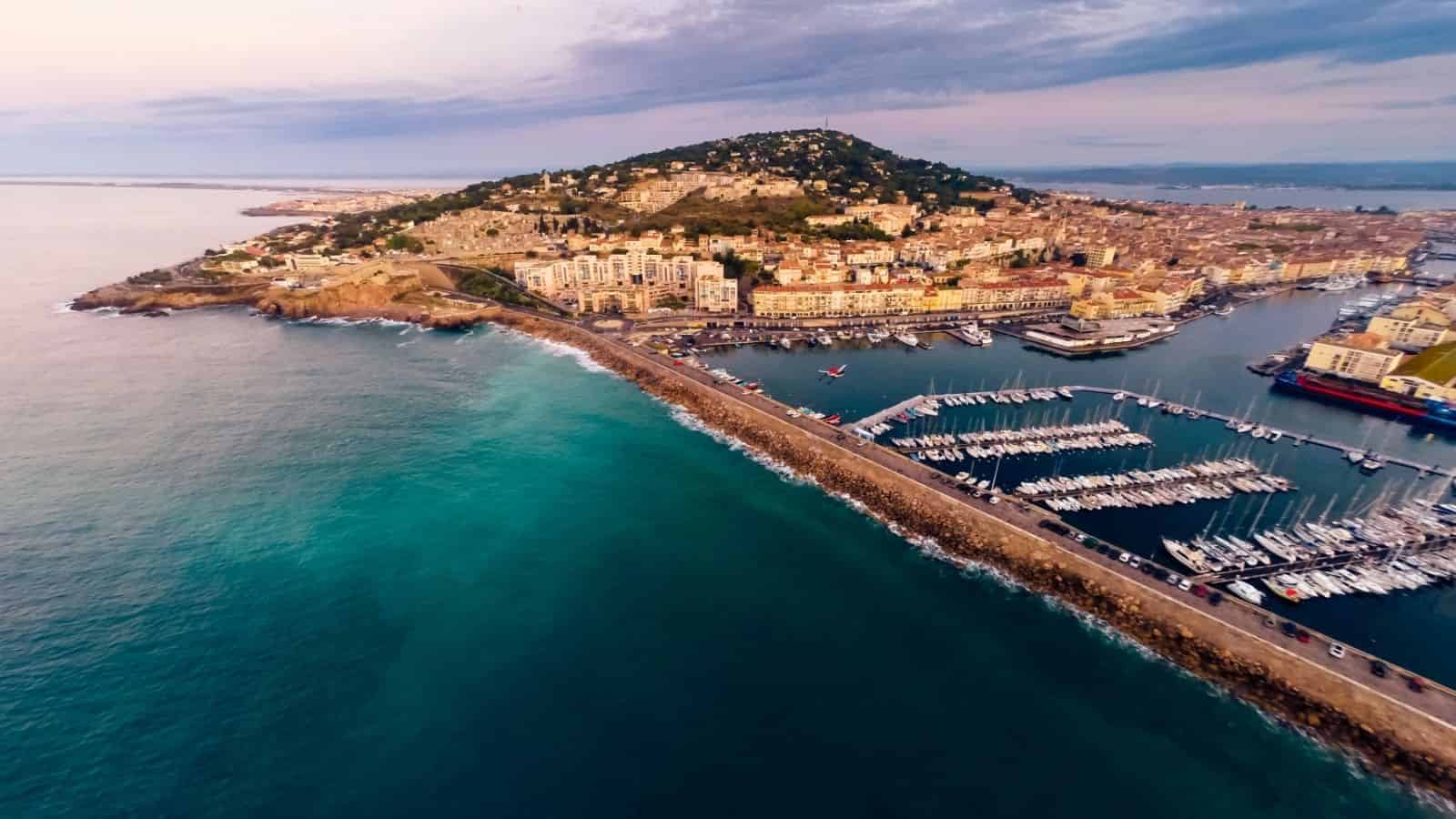 Aerial view of a coastal town with a marina, breakwater, and hillside houses by the sea.