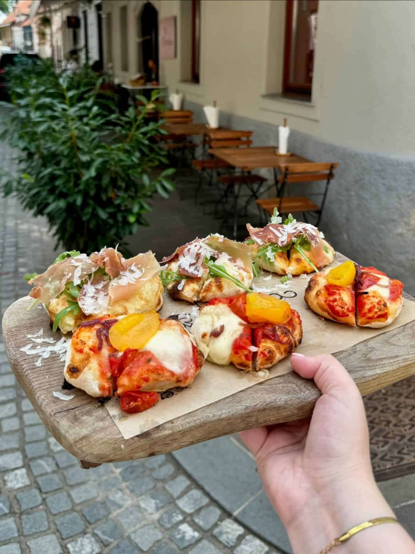 A hand holds a wooden board with assorted mini pizzas, perfect for sampling at one of the city's top foodie destinations, with outdoor café tables visible in the background.